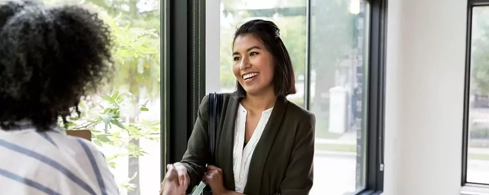 Two women shaking hands by a window, smiling at each other.