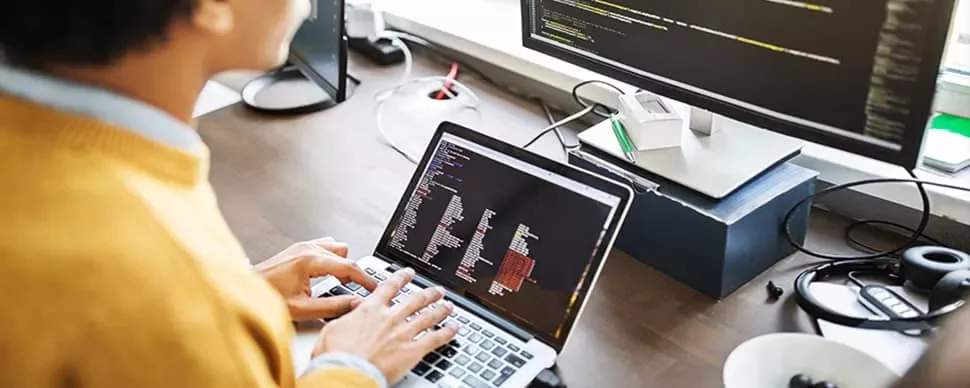 Person coding on a laptop with monitor showing code.