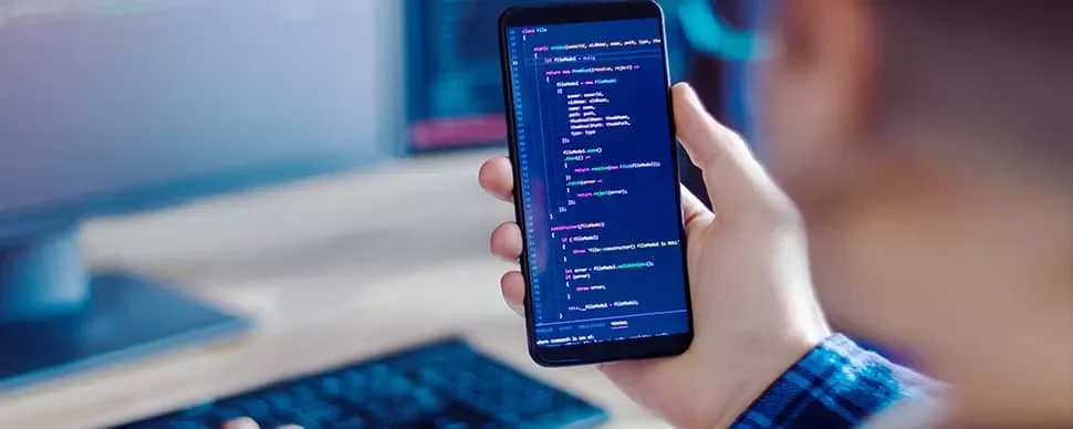 Person holding a smartphone displaying code, sitting at a desk with a computer monitor in the background.