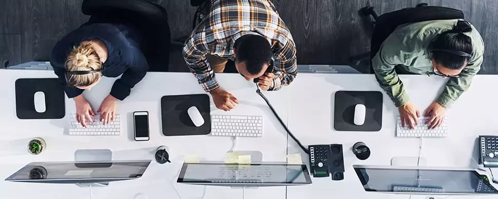 Top view of three people working at white desks with computers and headsets in an office.