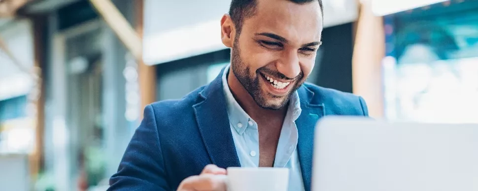 Man smiling while using a laptop in a cafe, holding a cup of coffee.