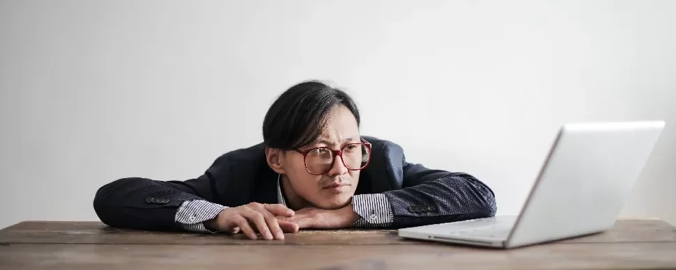 Person with glasses leaning on table, looking at laptop screen thoughtfully