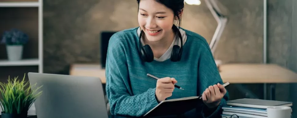 Smiling woman with headphones writing notes at a laptop.