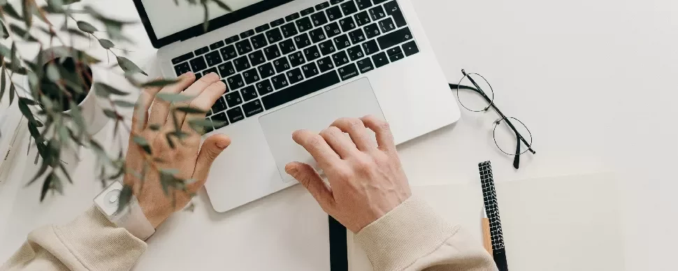 Person working on a laptop at a desk with glasses and a notebook nearby.