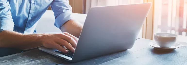 Person typing on a laptop next to a cup on a wooden table in sunlight.