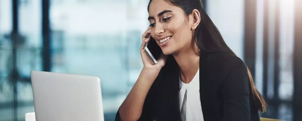 Woman smiling while talking on the phone and using a laptop in an office setting.