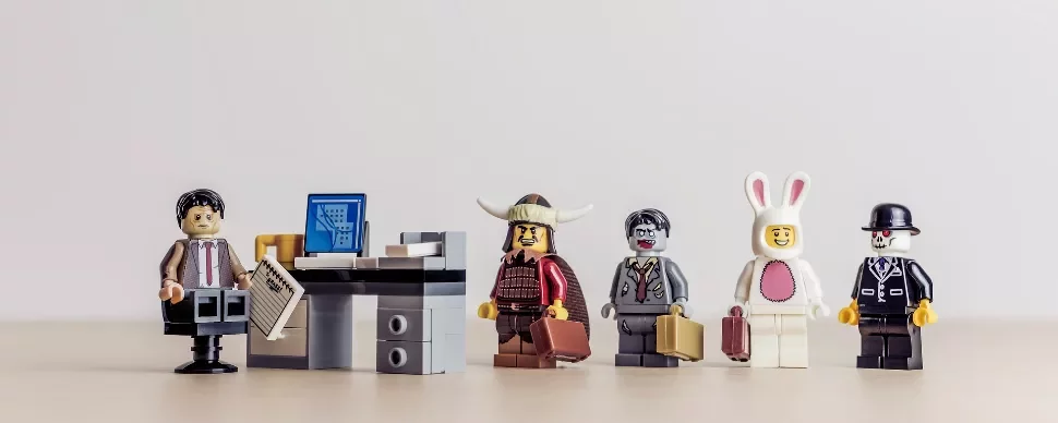 LEGO figures dressed in various costumes stand in line beside an office desk with a computer.