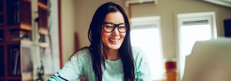 A woman with glasses smiling while using a laptop at home.
