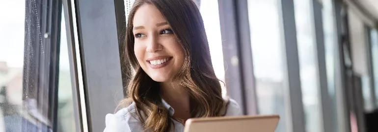 Smiling woman holding a tablet, looking out a window.