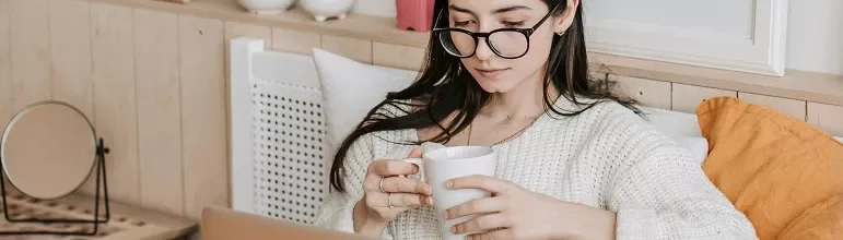 Person wearing glasses, sitting on a bed with a cup, and using a laptop.