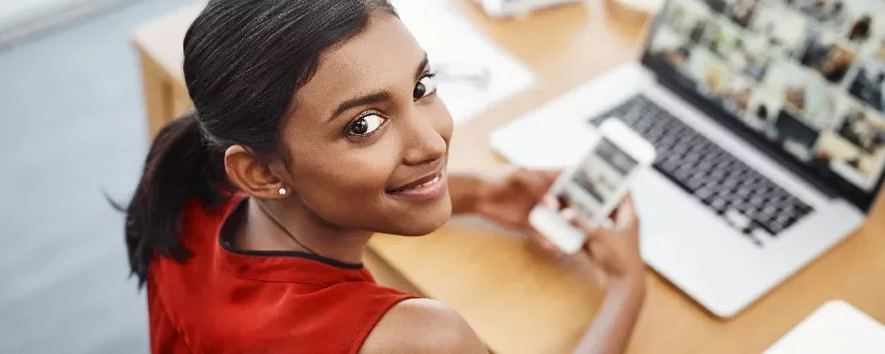 A female HR assistant sitting in front of an open laptop smiling and holding a mobile phone.
