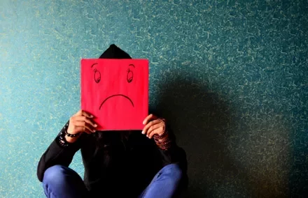 Person sitting against a wall holding a red sad face drawing over their head.
