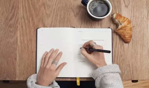 Person writing in a notebook with a coffee and croissant on a wooden table.