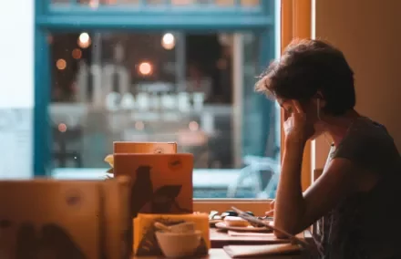 Person sitting in a cafe by a window, wearing earphones and holding their head in one hand.